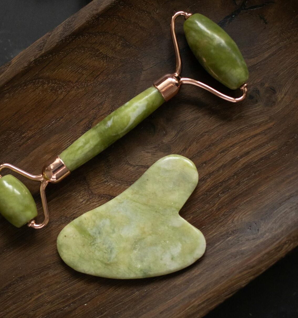 From above of Jade roller and Gua Sha massage tool placed on wooden plate on black marble table with fresh twigs of rosemary